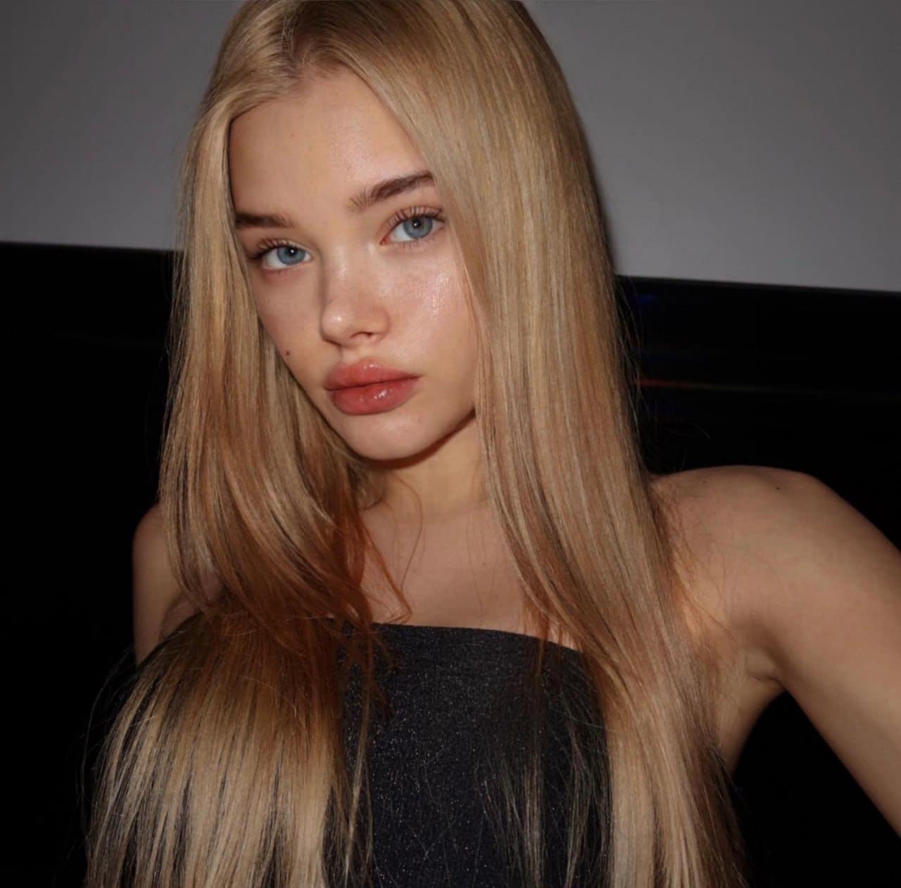 Close-up of a blonde woman in a black strapless top looking at the camera against a dark background.