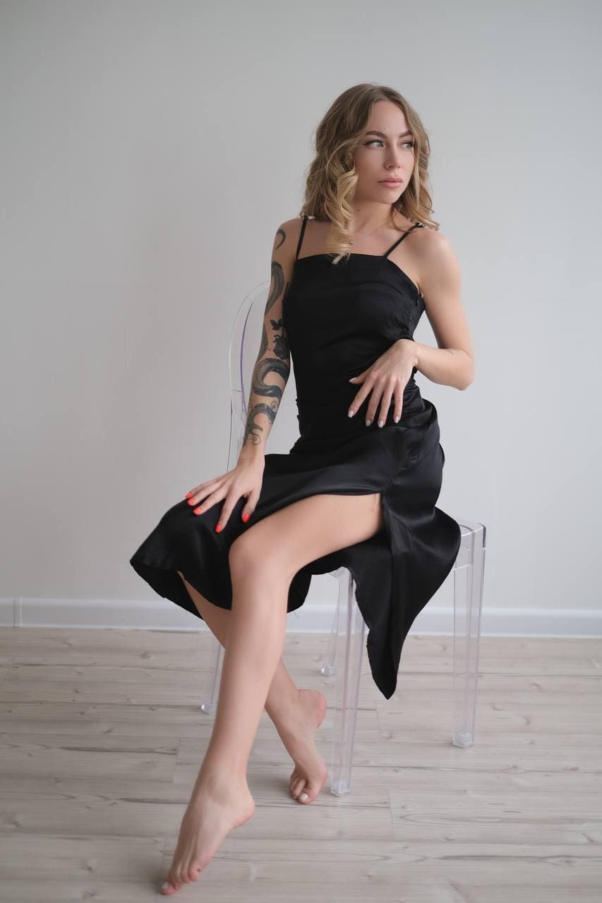 A woman sits barefoot on a clear chair in a black satin slip dress with a slit, against a plain wall and wood floor.