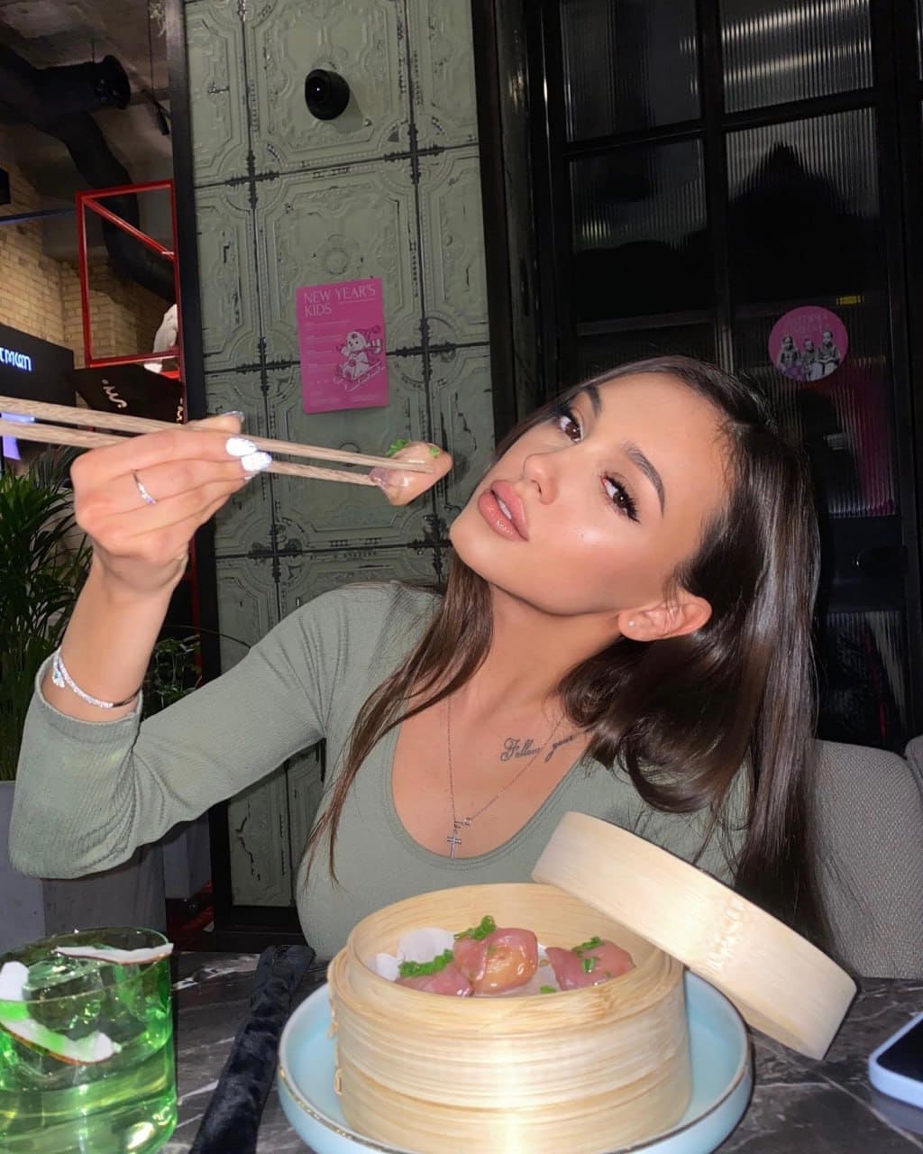 A woman in a sage-green top holds chopsticks with a dumpling at a restaurant table, with a bamboo steamer in front of her.