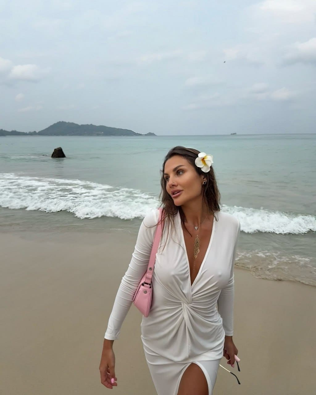 Dalila stands on a sandy beach in a white dress with a slit, holding sunglasses, with ocean waves and an island behind her.