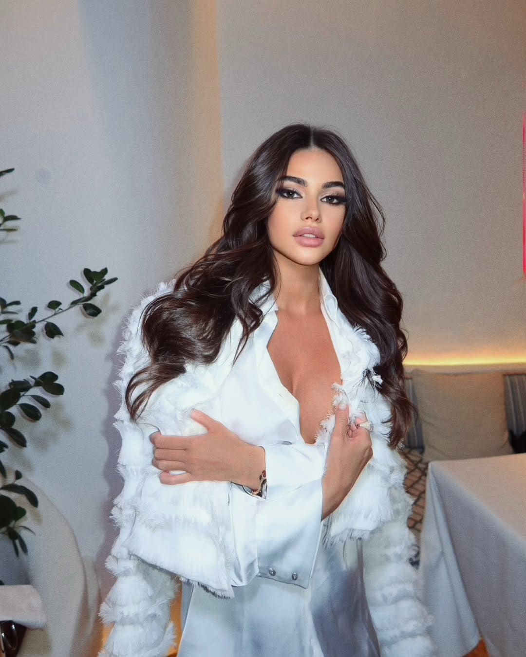 Indoors, she wears an open white satin shirt under a white feathered jacket, hand on chest, with a plant behind.