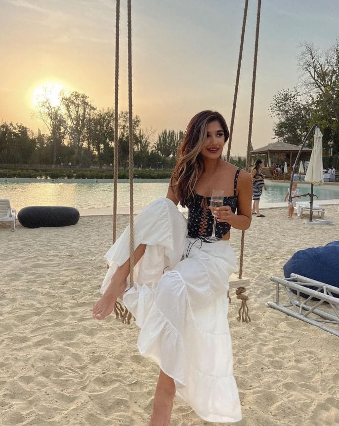 A woman sits on a rope swing by a pool at sunset, wearing a black crop top and white skirt while holding a glass of white wine.