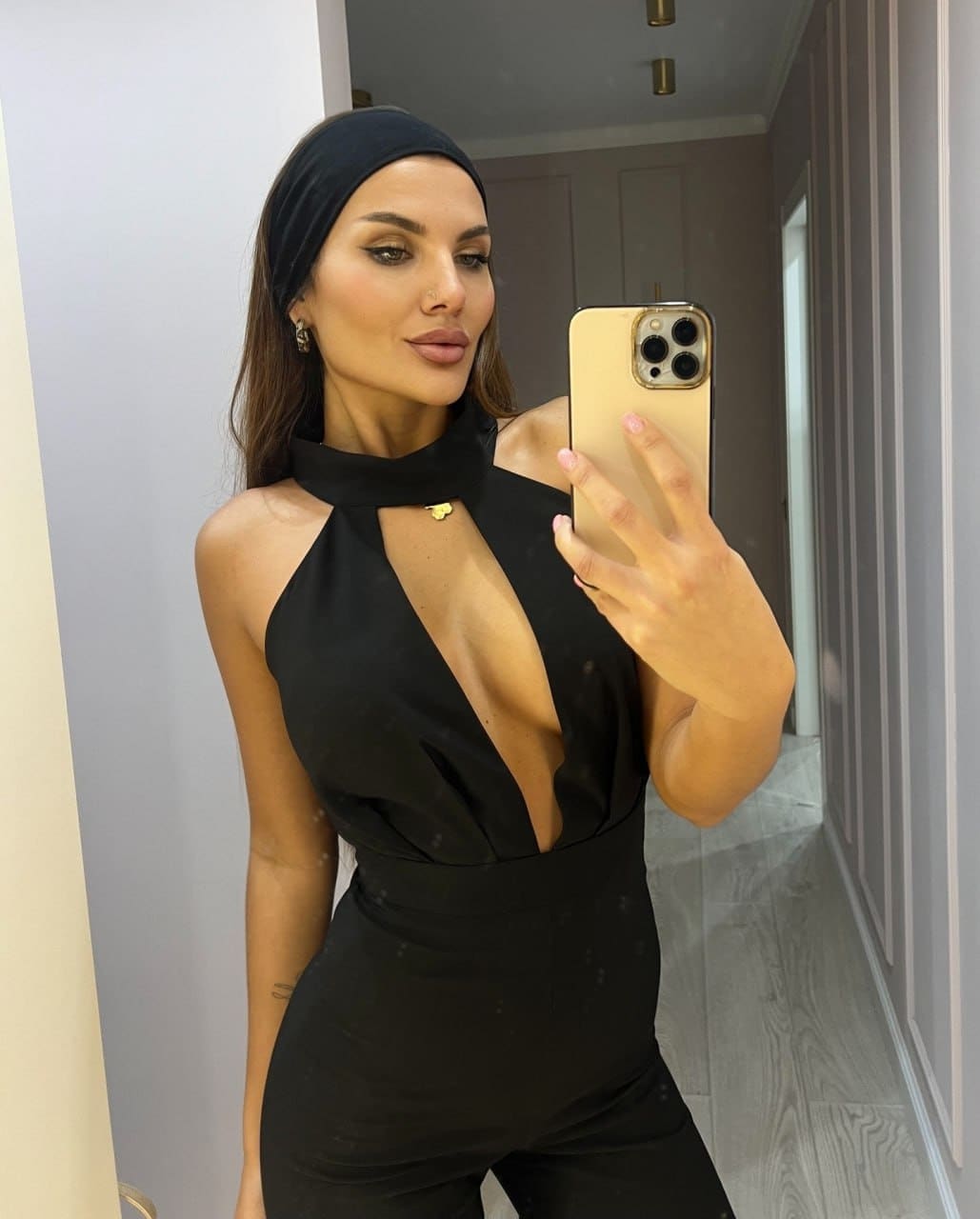 A woman with a black headband takes a mirror selfie in a hallway, wearing a black halter-neck dress with a plunging neckline.