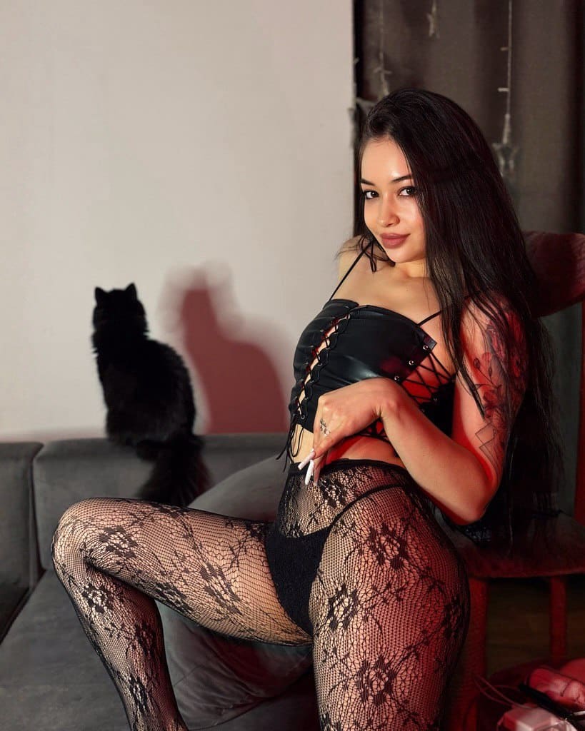 A woman sits on a gray couch in a black corset and floral fishnets, with a black cat sitting behind her.