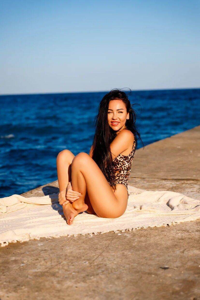 A woman sits on a pier towel in a leopard swimsuit, knees drawn up, hands resting on her feet.