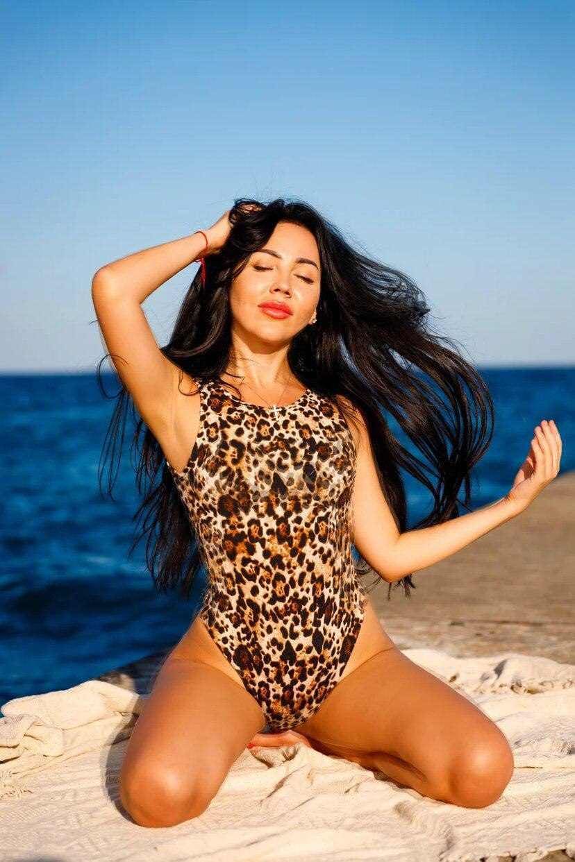 Eva kneels on a beach towel in a leopard-print swimsuit, one hand in her hair, against the sea.