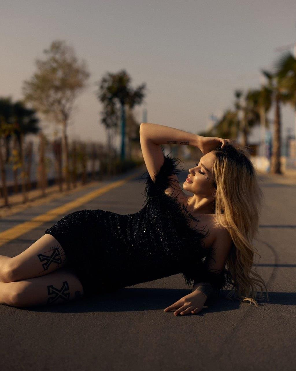 A woman reclines on asphalt in a black sequined dress with feathered sleeves, one arm behind her head, with palm trees in the background.