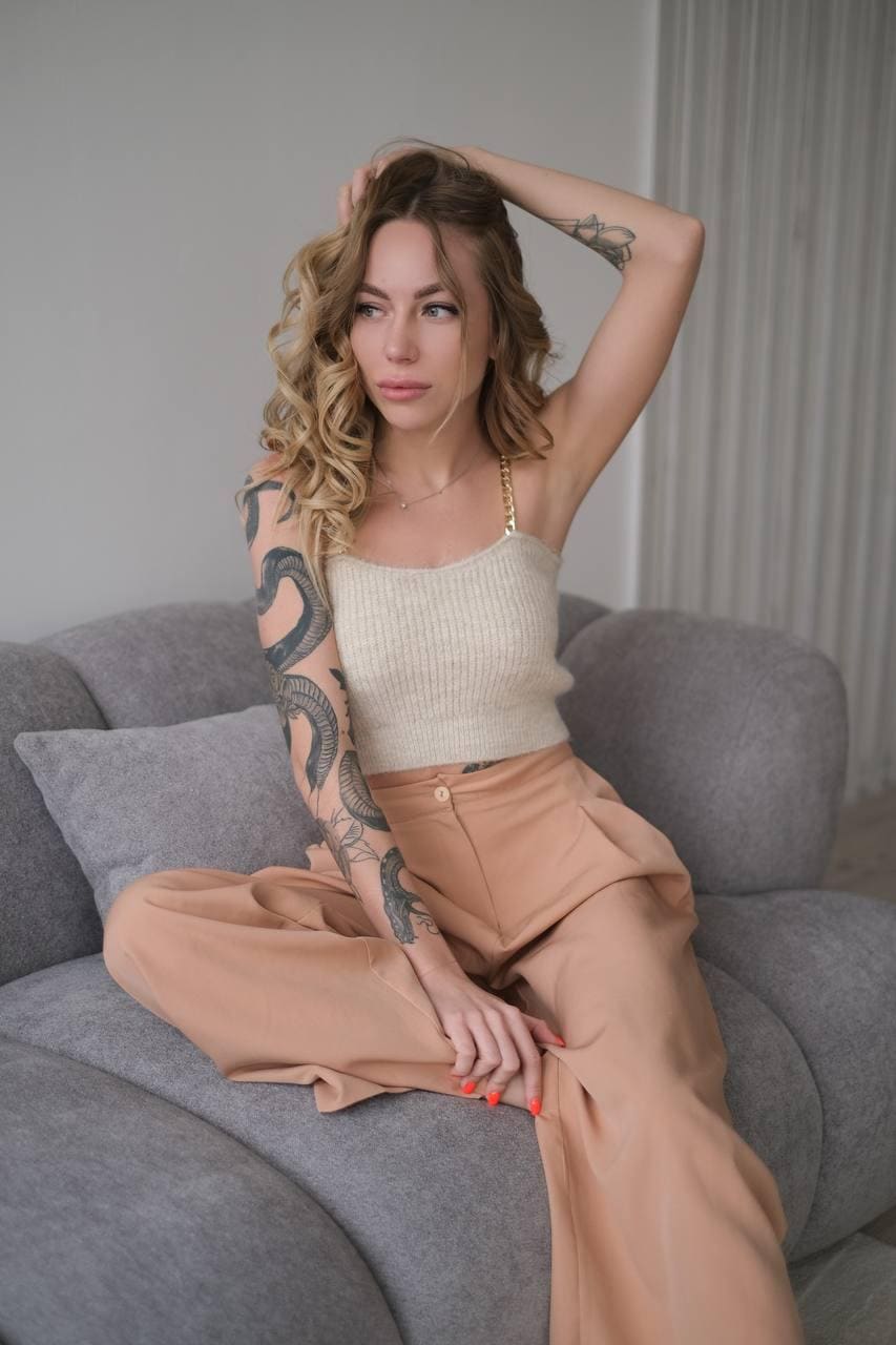 Jussie sits on a gray sofa in a cream camisole and tan trousers, one hand raised to her wavy blonde hair.