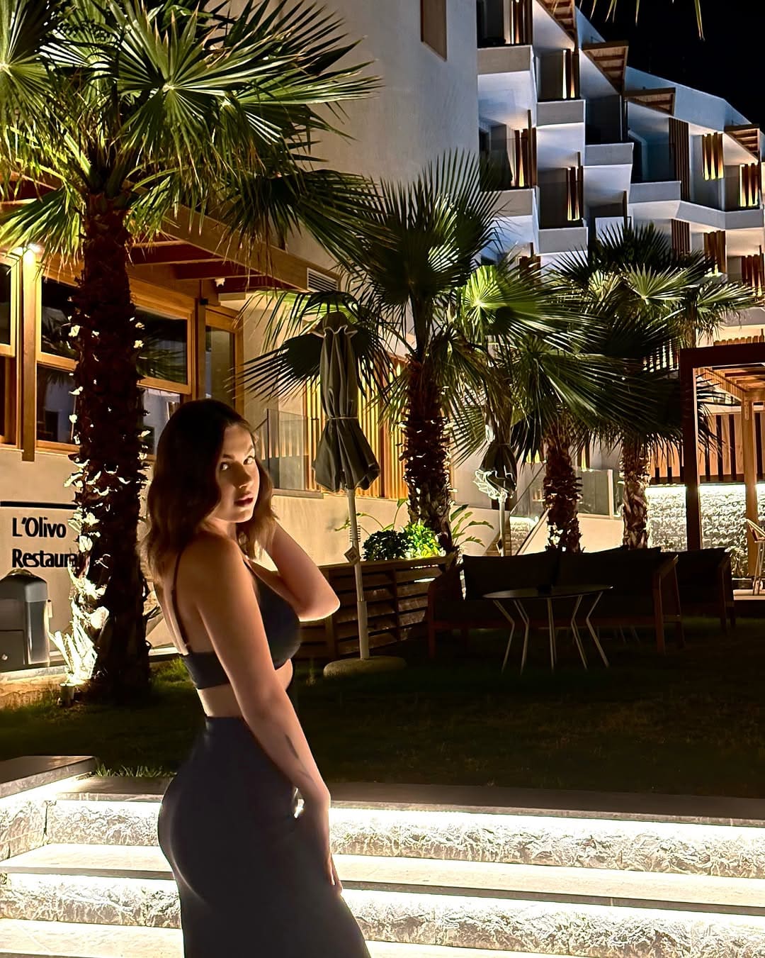A woman stands in profile on stone steps at night in a black bra top and long dark skirt, with palm trees and a lit restaurant behind her.