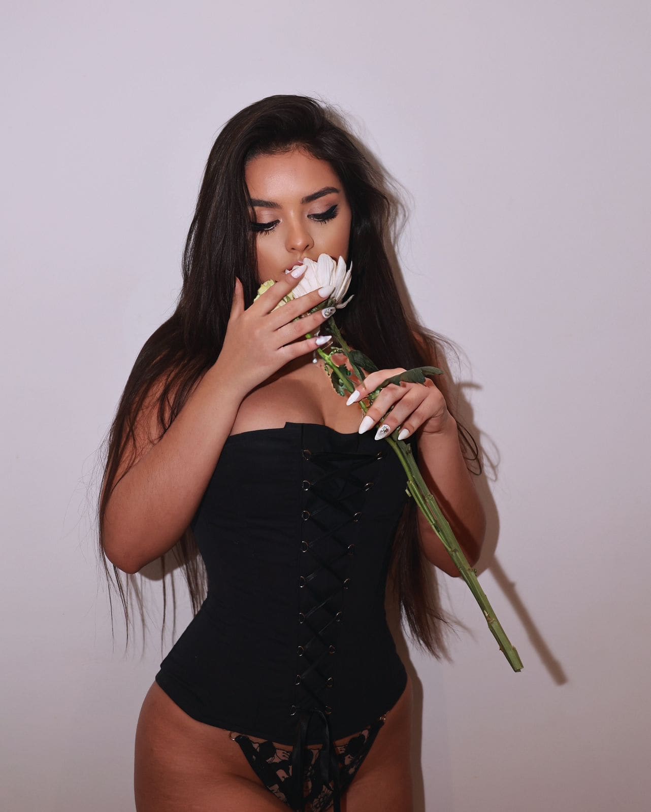 Against a plain light background, a woman in a black lace-up bodysuit holds a white flower near her face.