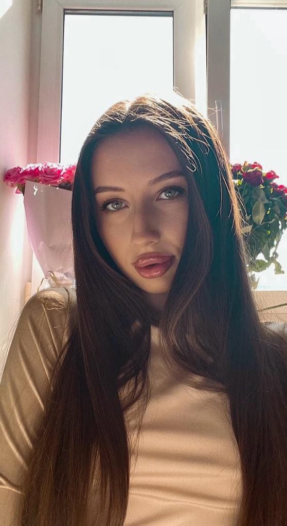 A woman sits indoors by a window, wearing a light top, with bouquets of pink and red flowers on the sill behind her.