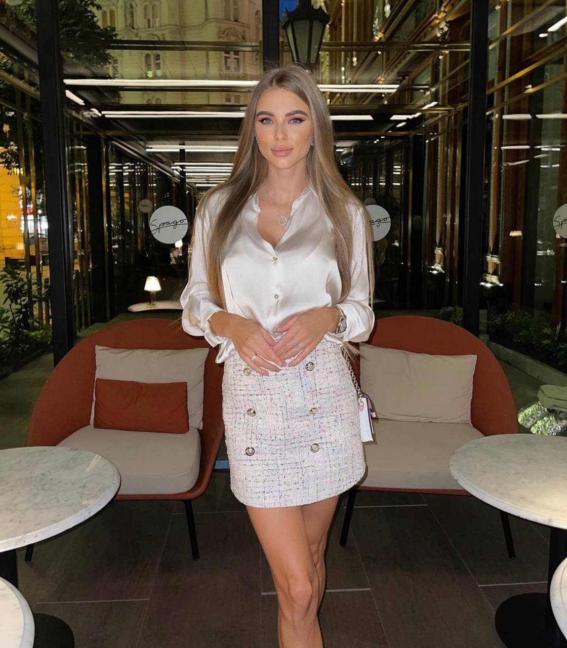 A woman in a white satin blouse and tweed skirt stands with hands clasped in a modern lounge featuring orange armchairs and marble tables.