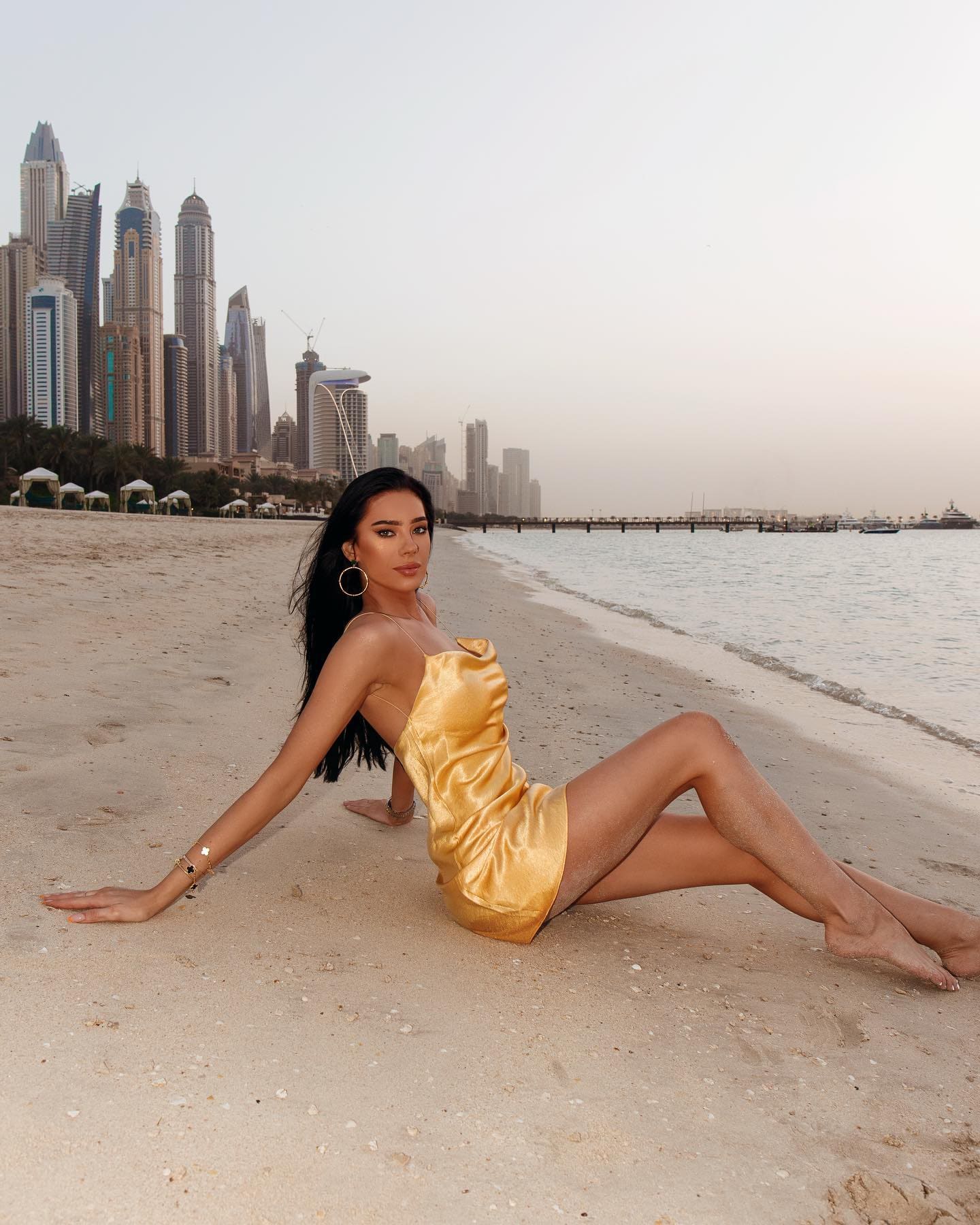A woman sits on sand in a short gold satin dress, legs extended and arm braced, with a city skyline in the background.