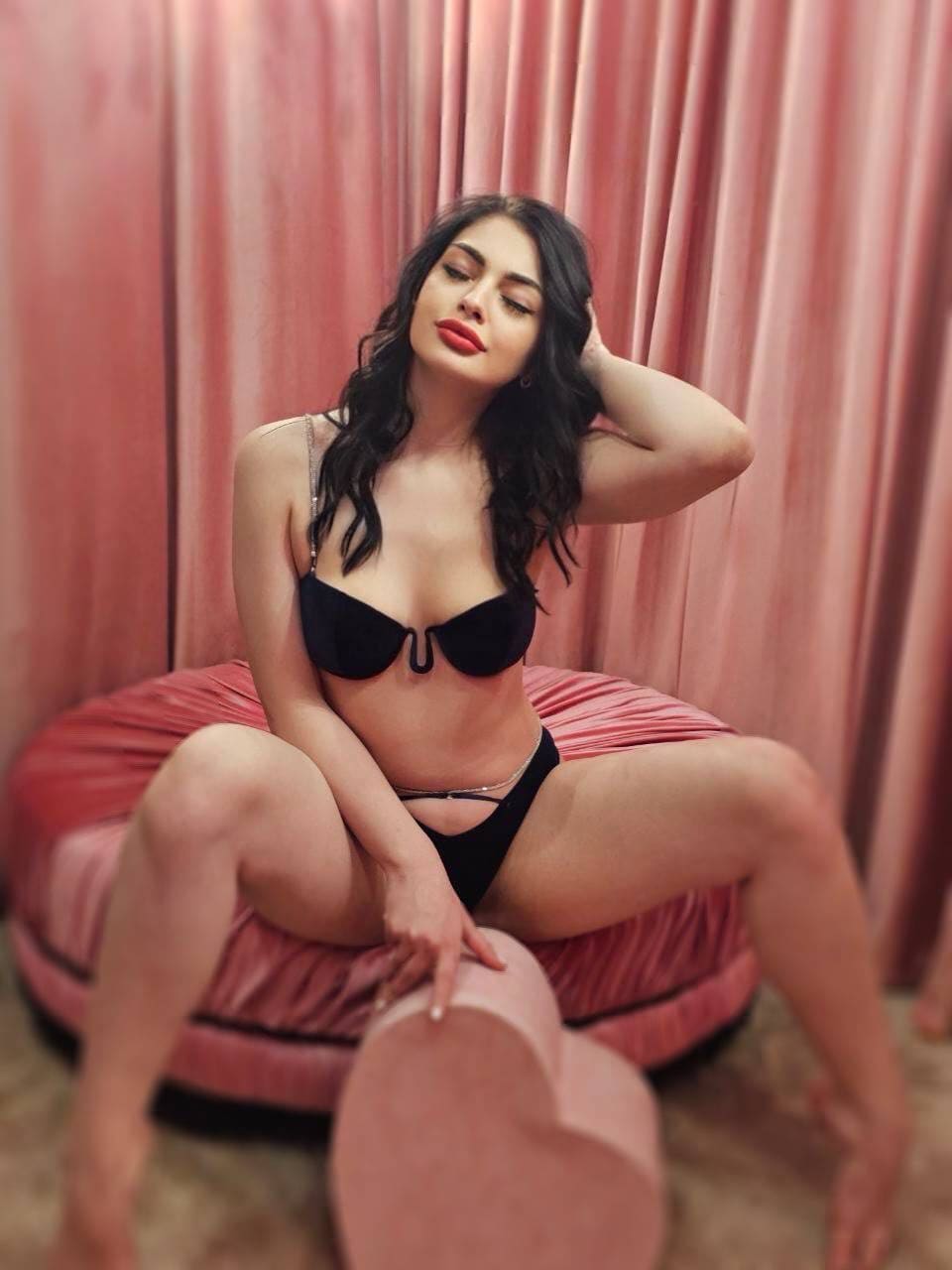 Seated on a round pink cushion against pink curtains, a woman in a black bra and underwear rests one hand on her head and the other on her knee.