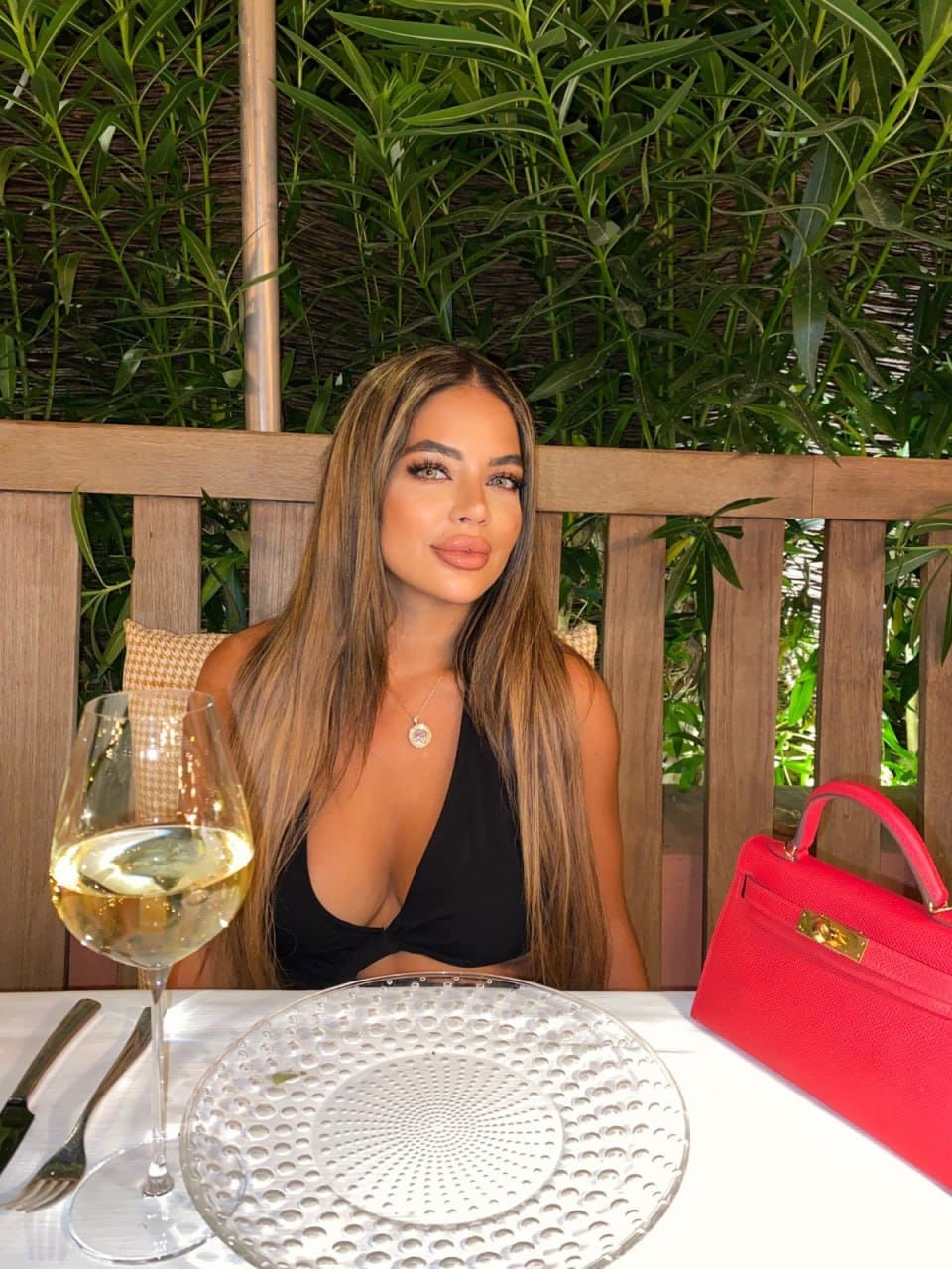 A woman sits at a white-clothed table outdoors, wearing a black deep-V top and pendant necklace, with a glass of white wine and a red handbag on the table.